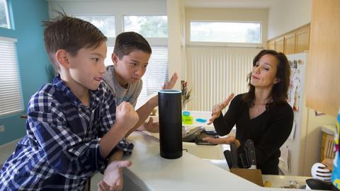 Zwei Kinder und eine Frau sitzen an einem Tisch. Auf dem Tisch steht das Gerät "Amazon Echo".