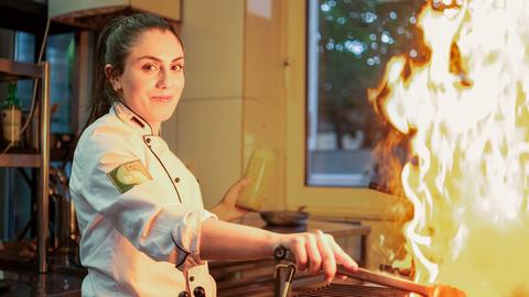 Eine junge Frau steht schmunzelnd vor einem Grillherd mit offener Flamme.