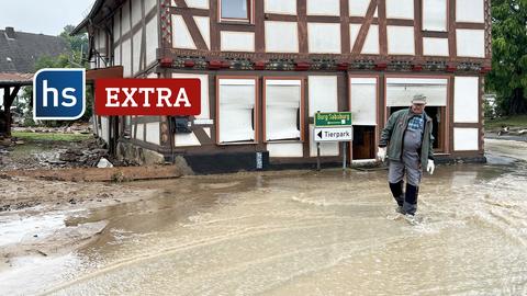 Gottsbüren/Trendelburg im Landkreis Kassel: Mit braunem Wasser überflutete Straße, die an einem Fachwerkhaus vorbeiführt. Vor dem Haus steht ein Mann mit Gummistiefeln knöcheltief im Wasser. Logo: hs extra