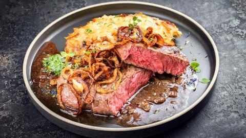 Traditioneller deutscher Zwiebelrostbraten aufgeschnitten mit Röstzwiebeln, brauner Sauce und Kartoffelgratin auf einem dunklen Teller.