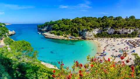 Das Bild zeigt den Ausblick über eine Bucht auf der Insel Mallorca. Das türkisfarbene Wasser mündet ins Meer. Am Strand sind einige Menschen mit Sonnenschirmen zu erkennen.