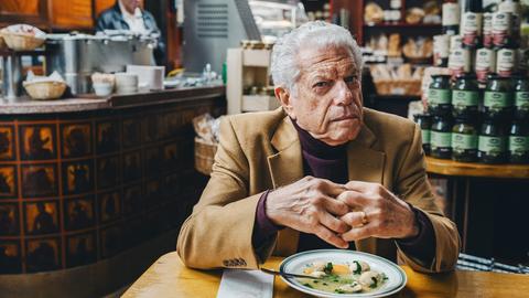 Symcha (Mike Burstyn) sitzt im Restaurant vor einem Teller Fischsuppe. 