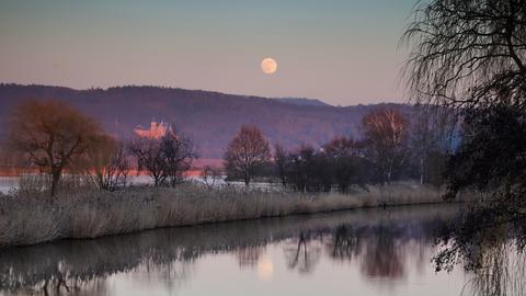 Ein Fluss im lilafarbem Abendlicht einem Schloss auf der gegenüberliegenden Seite und Vollmond am Himmel. 