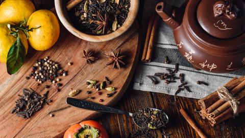 Zutaten für einen Masala Chai: Gewürze wie Anis, Zimt und Pfefferkörner auf Holzgrund. Daneben eine Teekanne.