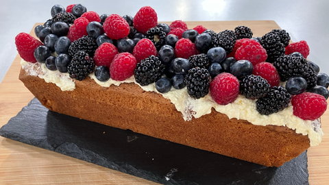 Joghurtfrischer Kastenkuchen mit beerigem Topping auf einer schwarzen, länglichen Schieferplatte.