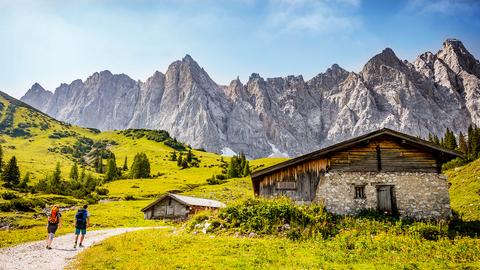 Zwei Wanderer laufen an einer Hütte vorbei mit Blick auf die Karwendel-Gebirgsgruppe.