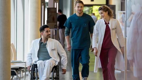 Arzt im Rollstuhl fährt neben Ärztin und Assistenzarzt den Gang entlang