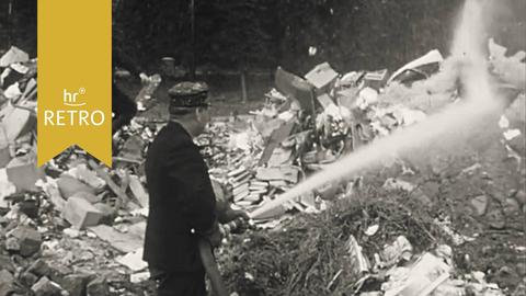 Ein Mann spritzt mit einem Feuerwehrschlauch auf brennenden Müll.