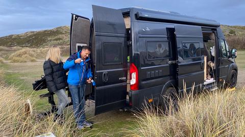 Ein Mann und eine Frau stehen in dicken Daunenjacken vor ihrem Campingwagen mitten in grasbewachsenen Dünen.